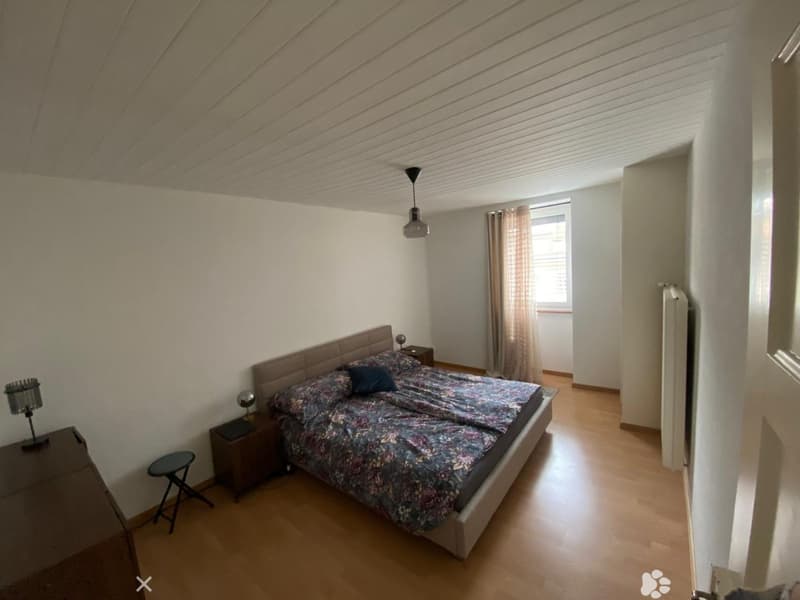 Bright bedroom with wooden flooring, a large bed with floral bedding, nightstands, a hanging lamp, and a window with curtains.
