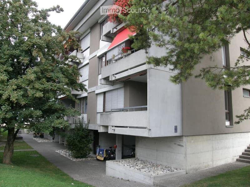 Modern multi-story apartment building with balconies, greenery, and a small parking area.