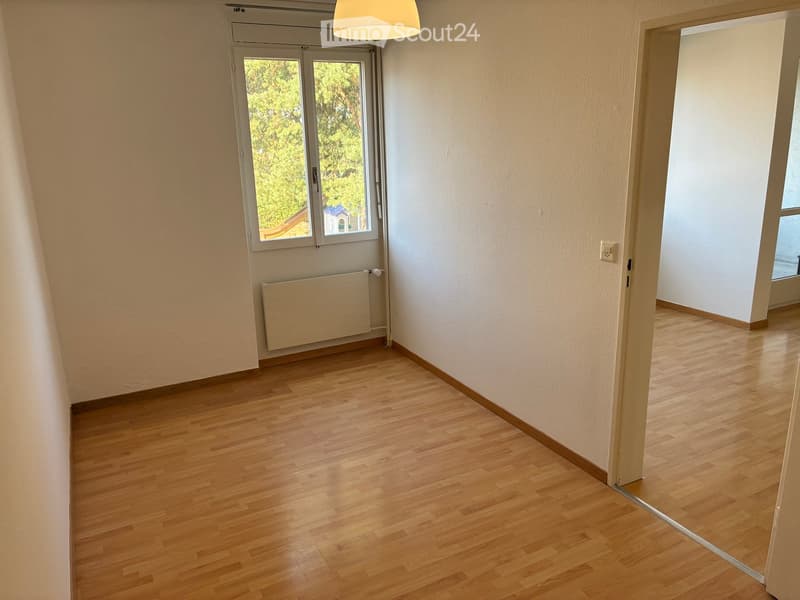 Empty hallway with wooden flooring, white walls, a radiator, and a window showing greenery outside.