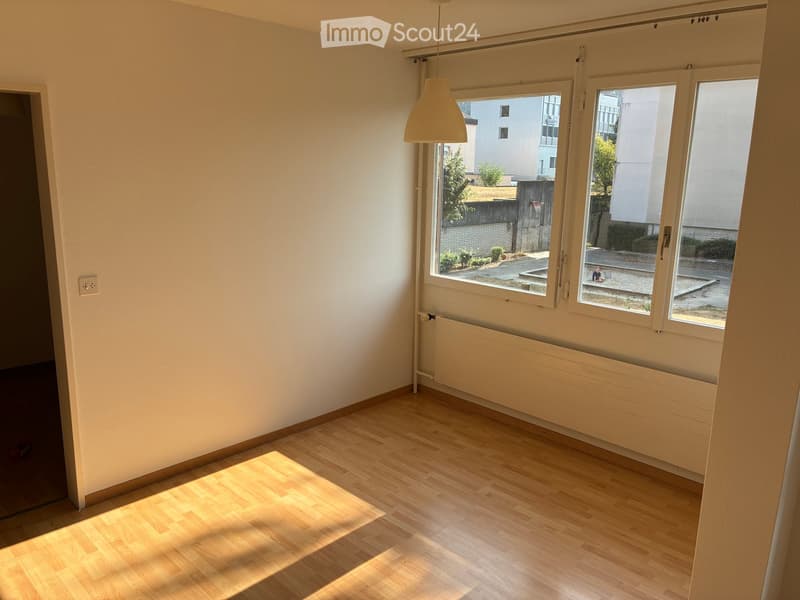 Empty room with wooden flooring, large windows, radiator, and a hanging light fixture.