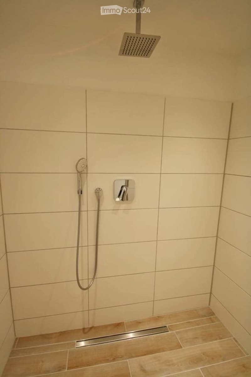 Modern shower area with beige tiled walls and floor, featuring a handheld shower hose, a wall-mounted shower control, and a fixed overhead showerhead.