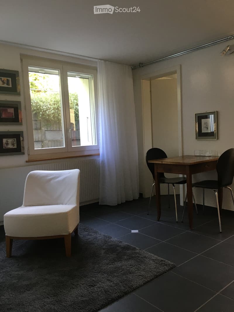 A modern, furnished room with a dark tiled floor, a white armchair, a wooden table with two black chairs, and a large window with white curtains. The walls are light-colored with several picture frames.