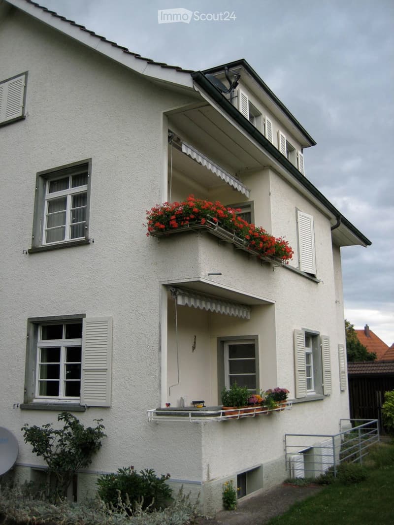 Two-story house with white exterior, multiple windows with shutters, balconies adorned with red flowers and potted plants, and a small garden area.
