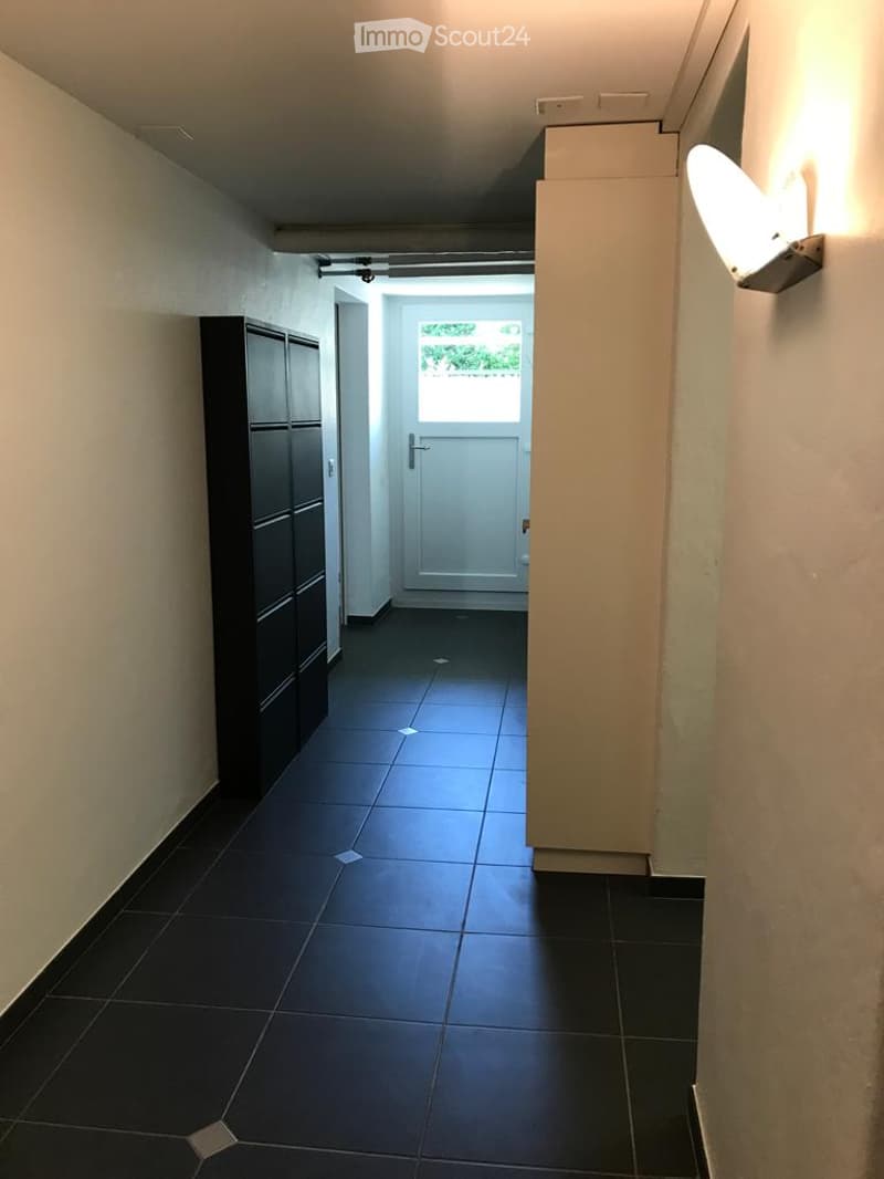 A narrow hallway with dark tiled flooring, light-colored walls, and a white door leading to an outdoor area. A tall black cabinet is on the left, and a wall-mounted light fixture is on the right.