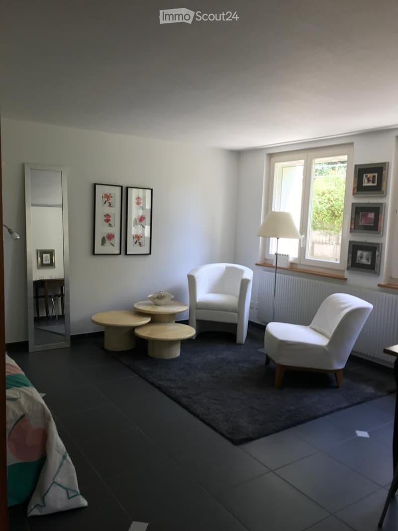 A modern, minimalist living space with white walls, dark flooring, and large windows. Features include a white armchair, small round tables, a floor lamp, and decorative picture frames.