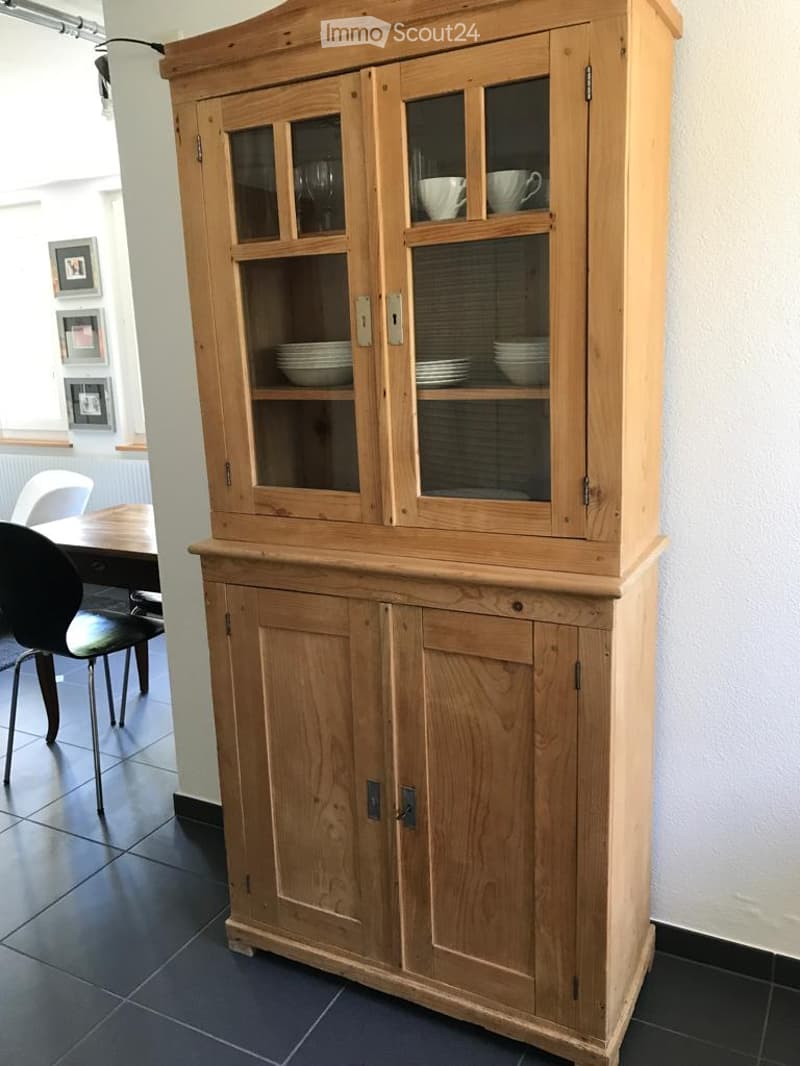 A wooden cabinet with glass doors displays dishes and cups. A black chair and a wooden table are visible in the background.