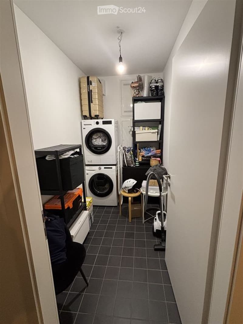 A small, well-organized laundry room with a washing machine, dryer, shelves, a small stool, and a fan. The floor is tiled, and the walls are white.