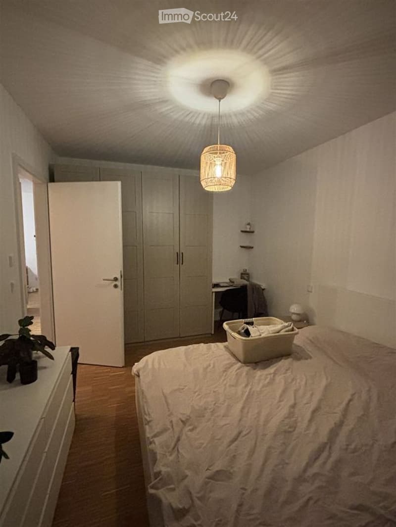 Cozy bedroom with a wooden floor, a large bed with white bedding, a wardrobe, a desk with a chair, a hanging woven lamp, and a small plant on a nightstand.