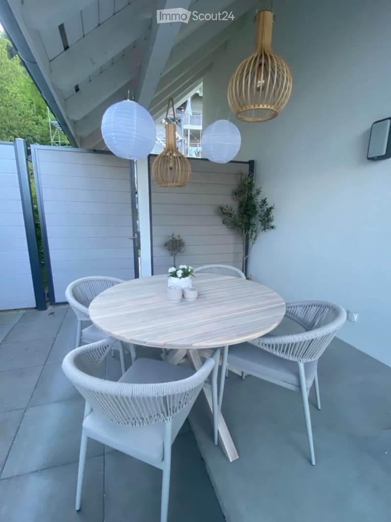 A modern outdoor patio with a round wooden table, four white wicker chairs, and hanging pendant lights. The area is enclosed with a white wooden fence and features small decorative plants.