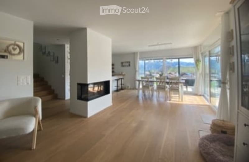 Bright, modern living and dining area with wooden flooring, large windows offering a scenic view, a fireplace, a sofa, and a dining set.