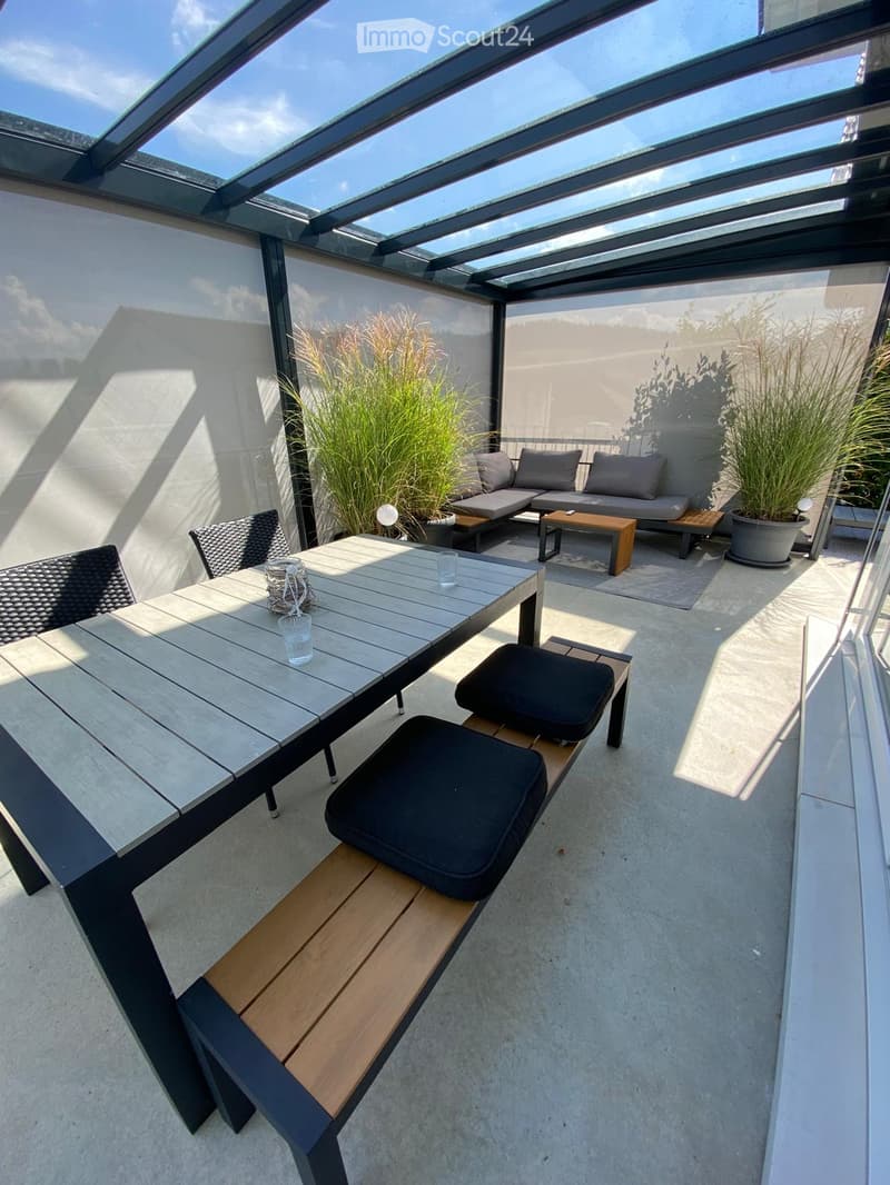 A modern, covered balcony with a glass roof, featuring a wooden table with black chairs, a sofa, a bench, and potted plants.