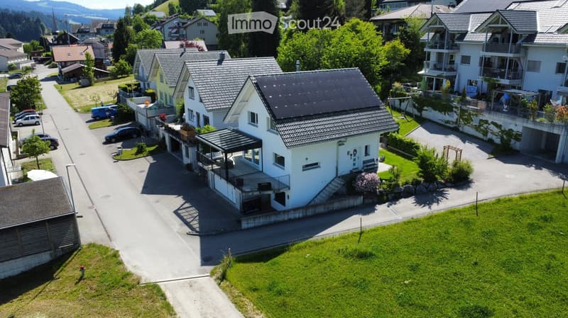 Aerial view of a residential area with multiple houses, driveways, and parked cars. The houses have varied architectural styles and are surrounded by greenery.