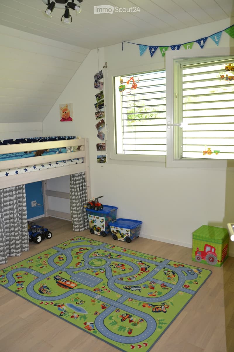 A children's bedroom with a bunk bed, a road-themed play mat, toy cars, a toy tractor, storage bins, and a green stool. The room features wooden flooring, white walls, and large windows with blinds.
