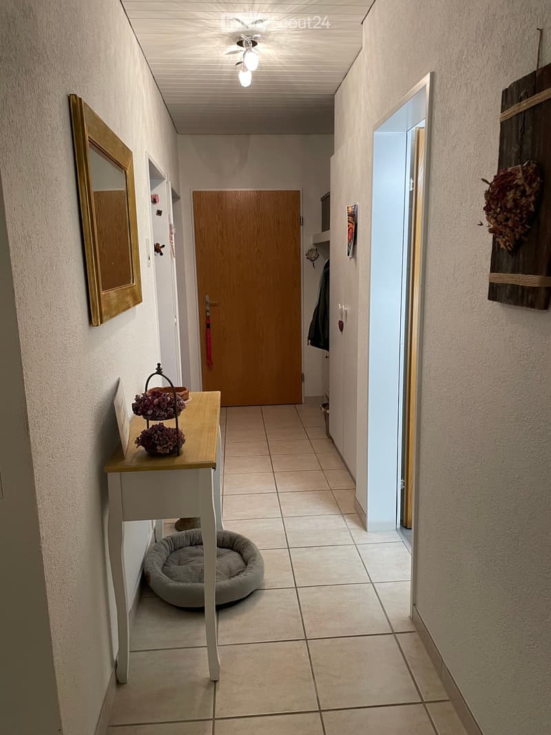 A well-lit hallway with tiled flooring, a small wooden console table with floral decorations, a pet bed, and a coat rack with a coat hanging on it. The hallway leads to a wooden door and has a ceiling light fixture.