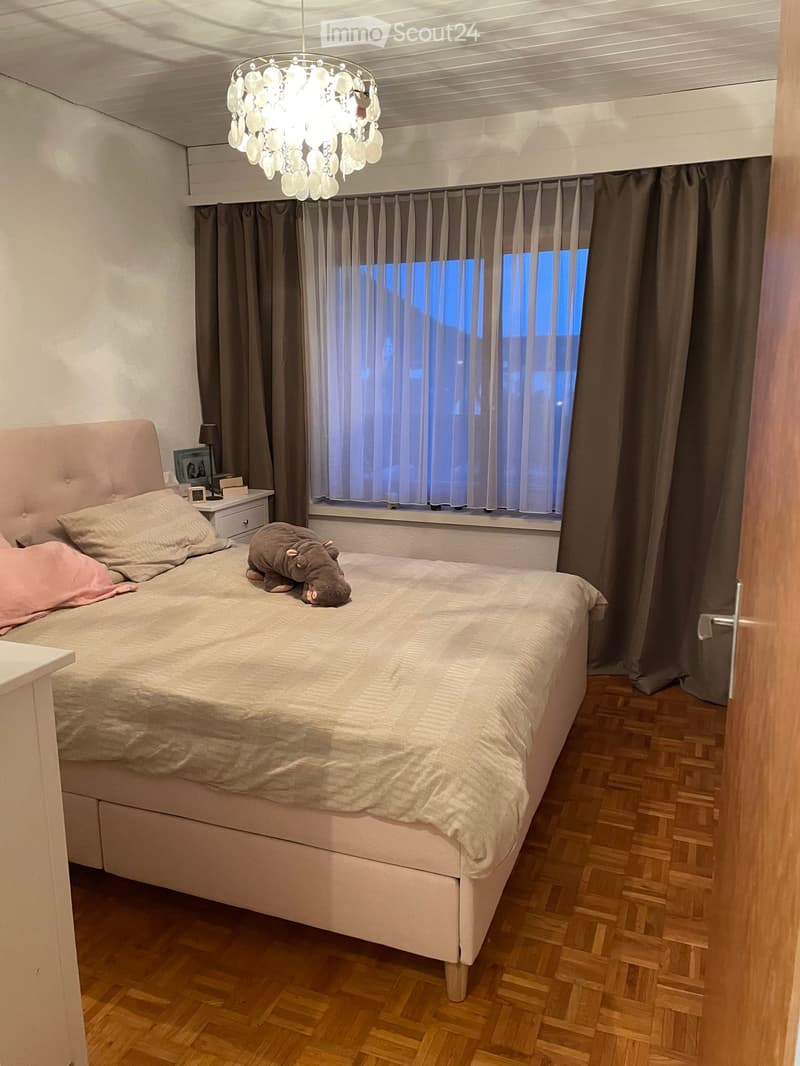 Cozy bedroom with a large bed, nightstand, lamp, picture frame, and a stuffed animal on the bed. Wooden flooring and large window with curtains.