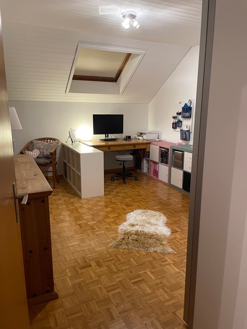 Cozy attic room with wooden flooring, slanted ceiling, and skylight. Features a desk with a computer, a wicker chair, a wooden bench, and a furry rug on the floor.