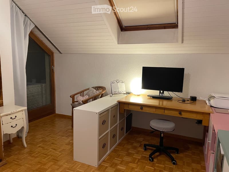 A well-lit attic room with wooden flooring, a large desk with a computer setup, a comfortable chair, a small cabinet, and a cozy seating area with a wooden chair and cushions.