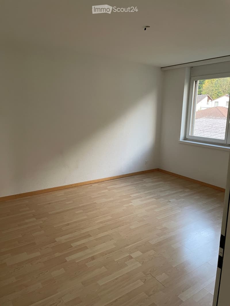 Empty room with light wooden flooring and white walls, featuring a large window with a view of a neighboring house and garden.