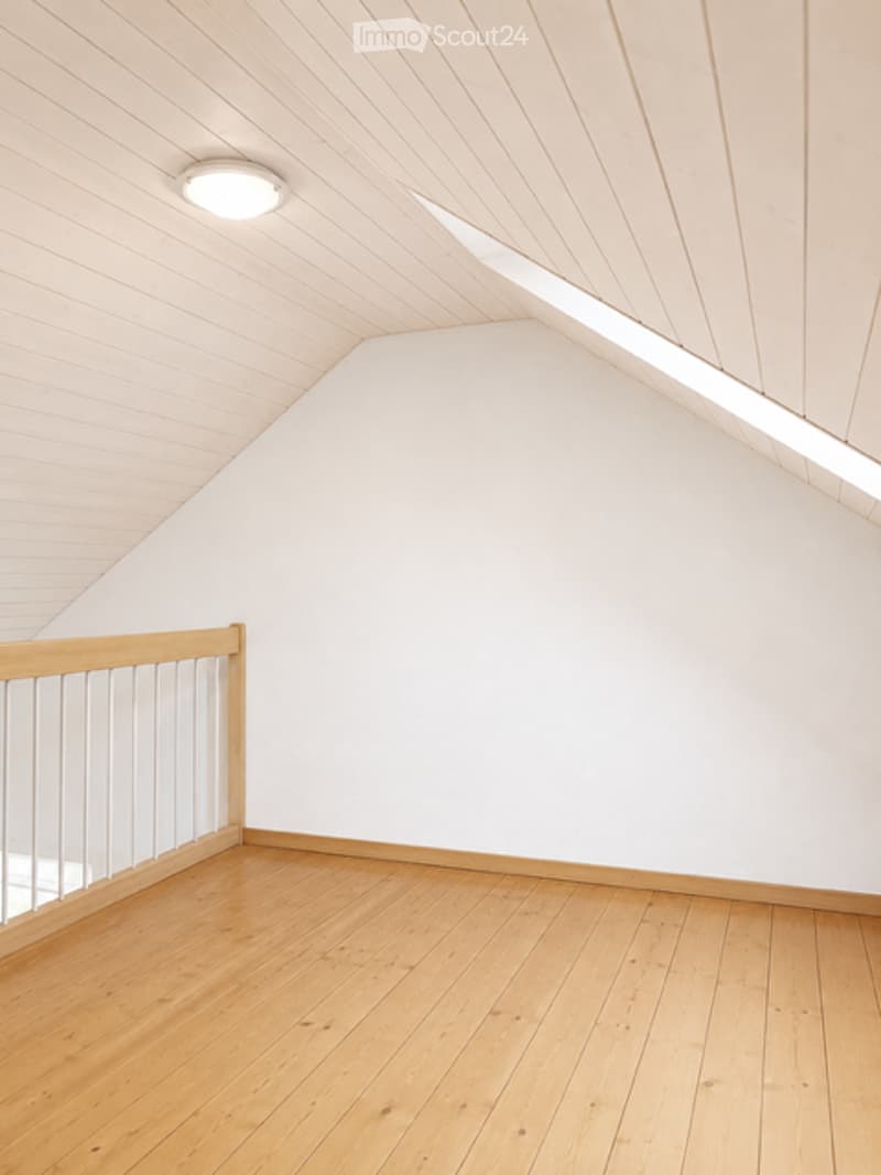 Empty attic room with wooden flooring, white walls, and a slanted ceiling with exposed wooden beams. A small railing is visible on one side.