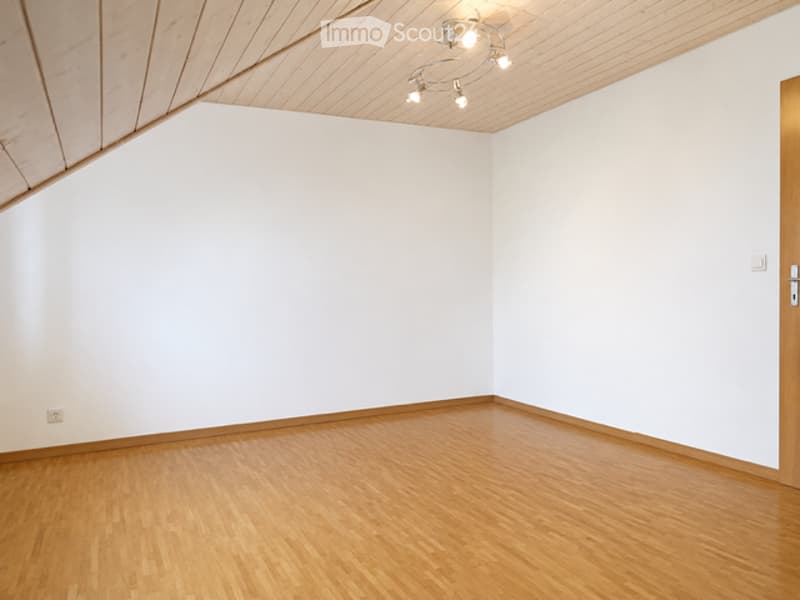 Completely empty room with white walls, wooden floor, and a slanted wooden ceiling with built-in lighting fixtures.