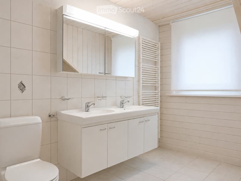 Modern bathroom with a white tiled wall, a double sink vanity with mirrored cabinet, and a toilet. The space features light wood paneling on the walls and a window with blinds.