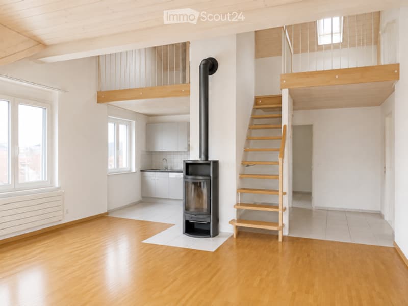 Bright, open-plan living space with wooden flooring, white walls, large windows, a staircase leading to an upper level, and a modern black wood-burning stove with a chimney.