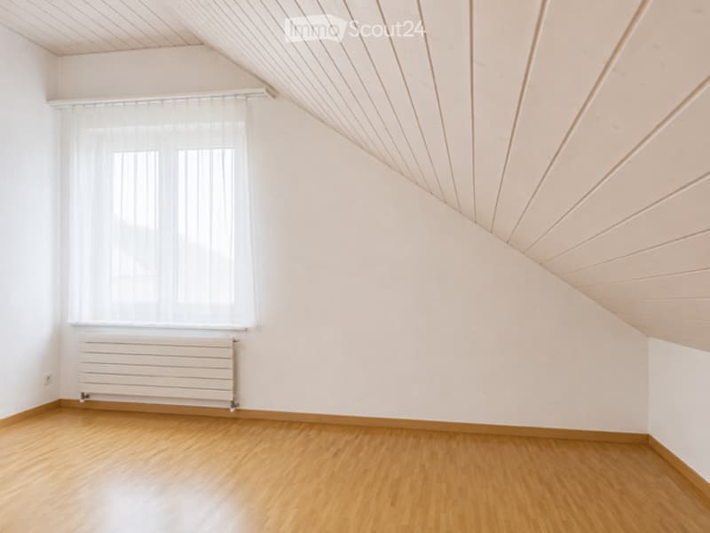 Empty attic room with wooden flooring, white walls, and a large window with white blinds. The ceiling has exposed wooden beams, and there is a white radiator below the window.
