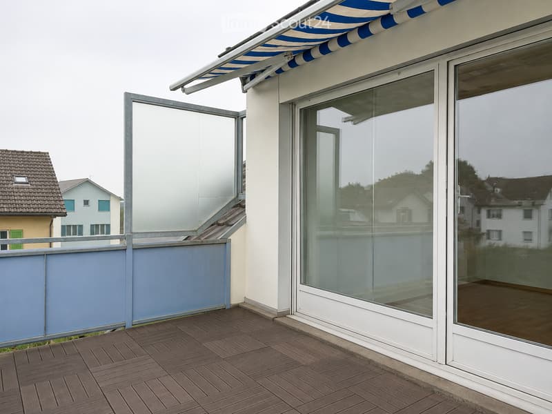 Balcony with wooden decking, glass sliding doors, and a retractable awning. Blue railing and modern design.