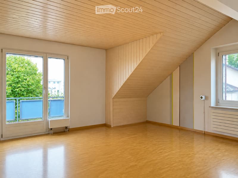 Completely empty room with wooden flooring, white walls, and large windows with blue blinds. The ceiling has a slanted design with wooden paneling.