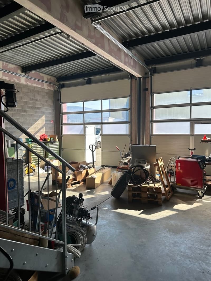 Empty garage with large roll-up doors, metal ceiling beams, and scattered storage items including boxes, tools, and equipment.