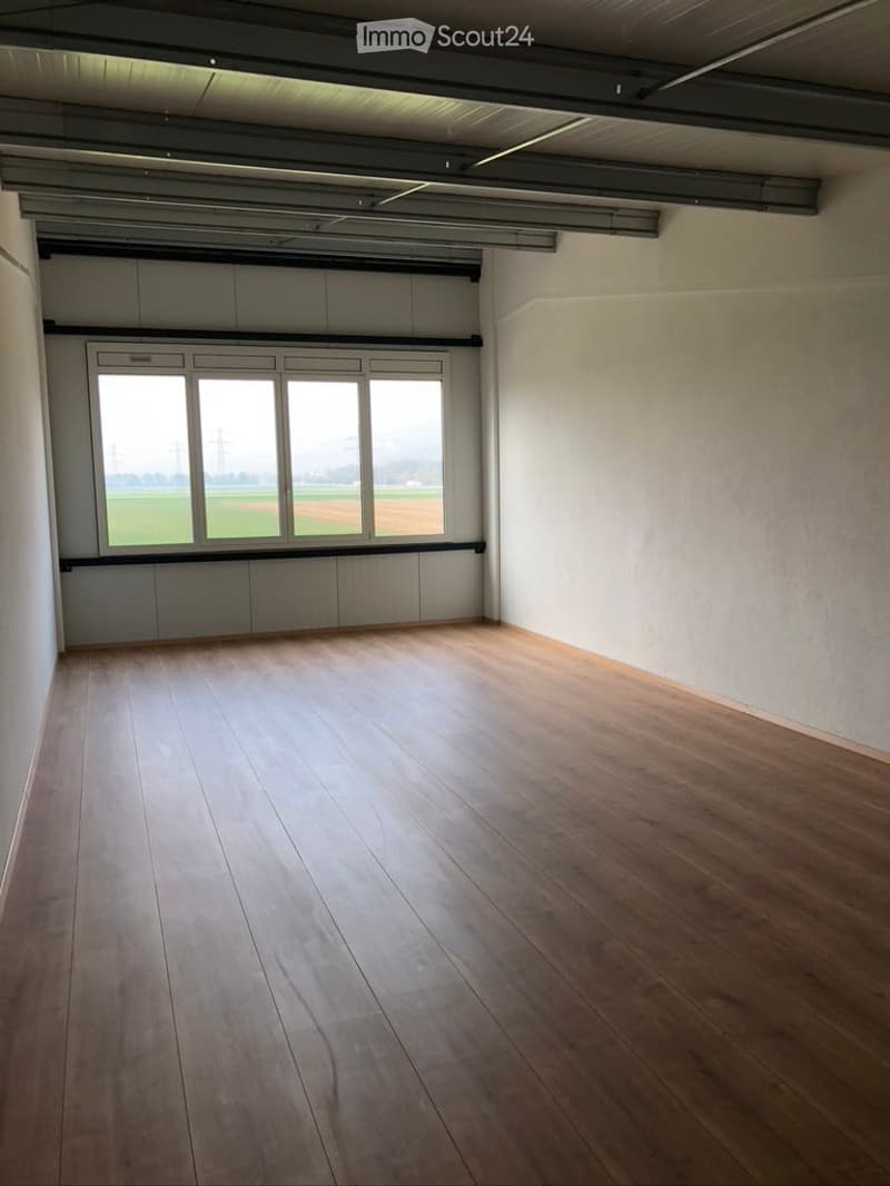 Empty room with wooden flooring, white walls, and large windows overlooking a field.