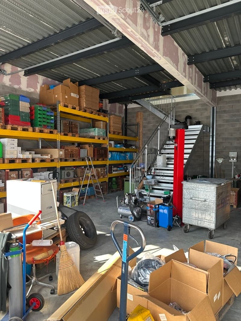 A spacious garage with metal shelving units filled with boxes and storage items, a staircase leading to an upper level, a refrigerator, a handcart, a broom, a tire, a compressor, and various other tools and equipment.