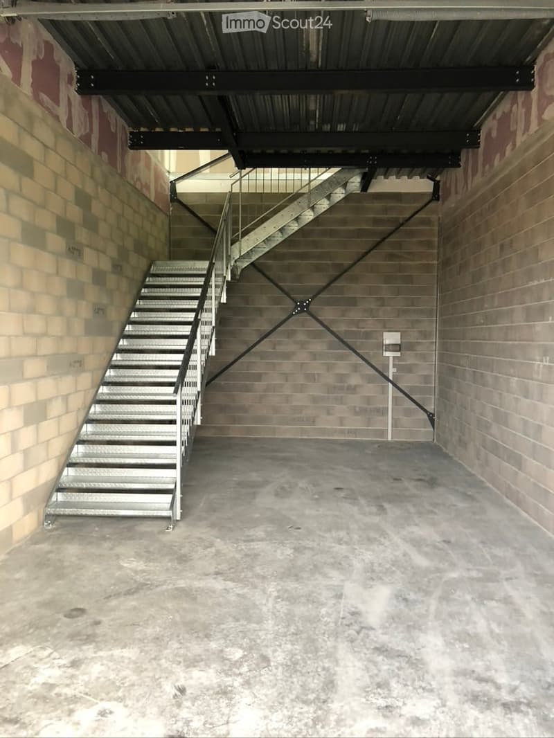 Empty concrete garage with a metal staircase leading to an upper level. The space has exposed brick walls and a metal ceiling structure.