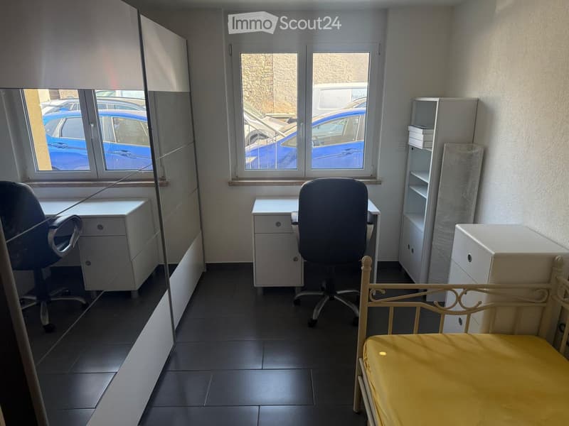 A furnished room with a bed, desk, chair, and cabinet. The room has large windows with blinds and a view of parked cars outside.