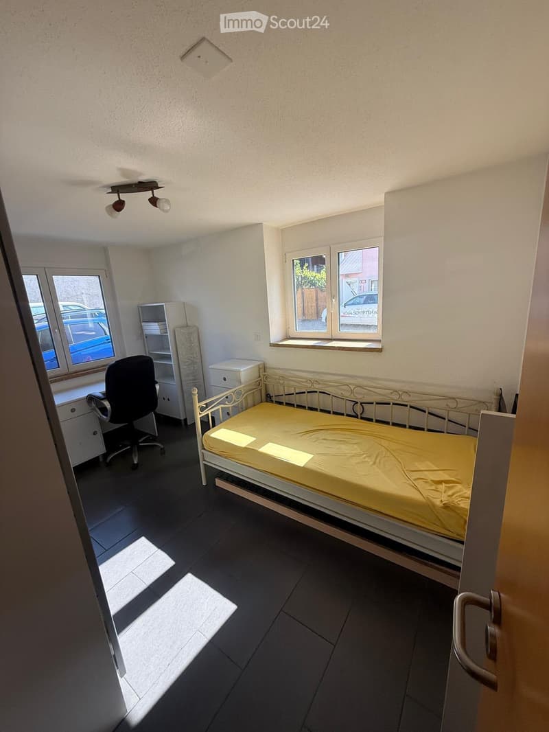 A small bedroom with a single bed, a desk with a chair, and a cabinet. The room has dark tiled flooring and white walls, with natural light coming through two windows.