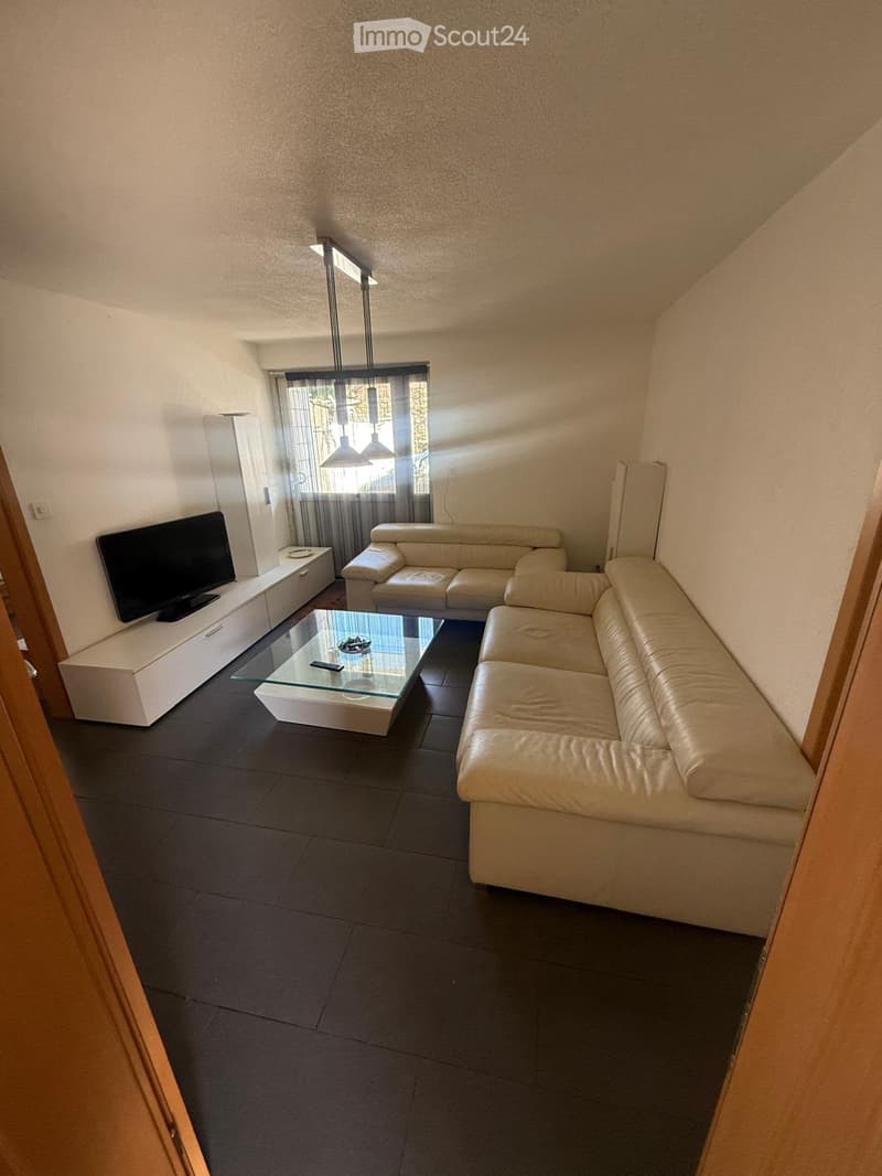 Modern living room with a white leather sofa, glass coffee table, flat-screen TV on a white stand, and pendant lighting over the window.