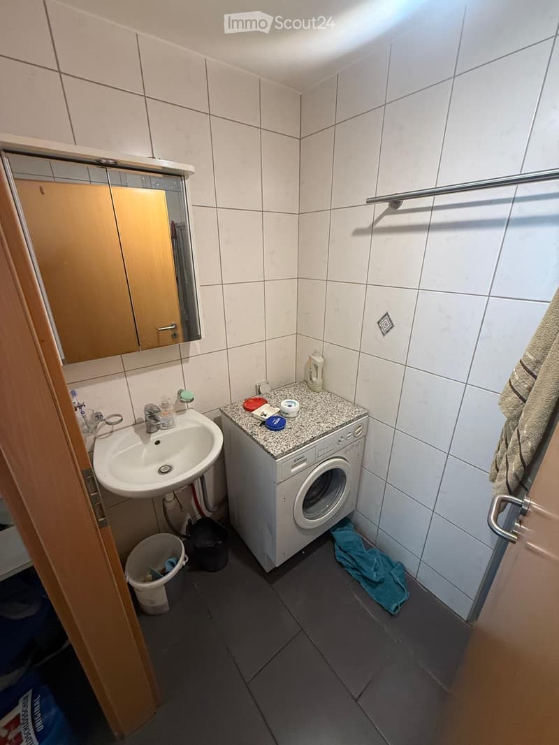 Small bathroom with a sink, mirror, washing machine, and towel visible. The walls are tiled, and the floor is dark grey.