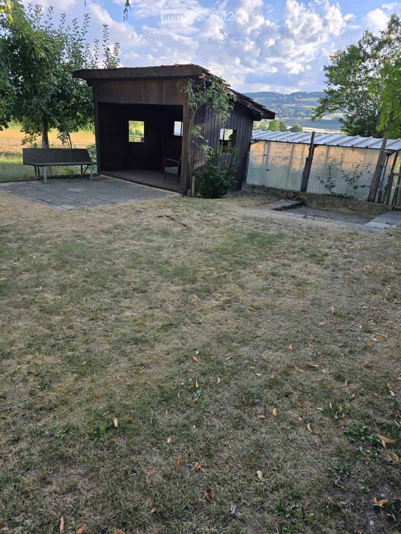 A small wooden outdoor structure with a bench and a chair, surrounded by grass and trees, with a view of a field and hills in the background.