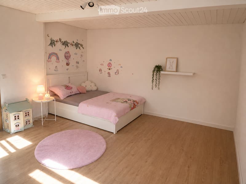 Cozy children's bedroom with a white bed, pink bedding, a round pink rug, a small nightstand with a lamp, a toy house, and wall decals featuring clouds and rainbows.