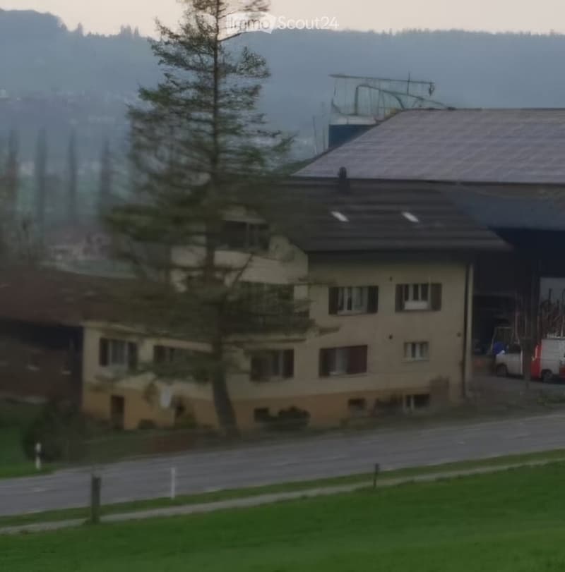 A multi-story residential building with a sloped roof, multiple windows, and a visible garage entrance. The building is surrounded by a grassy area and trees, with a road in front.