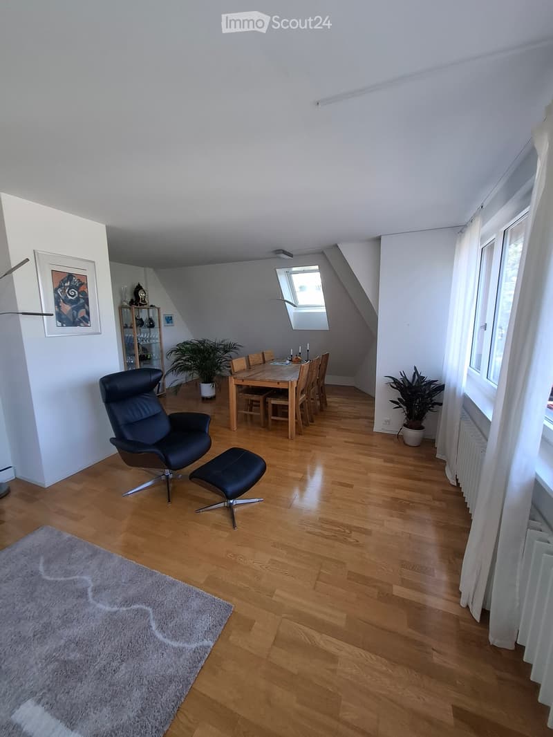 Open-plan living and dining area with wooden flooring, a black leather chair and stool, a dining table with chairs, a cabinet, and plants.
