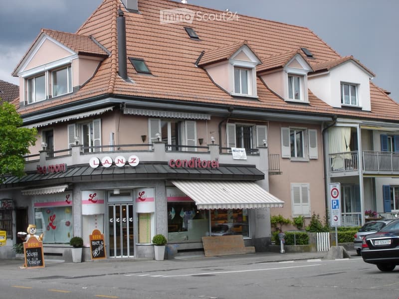 Two-story building with a tiled roof, multiple windows, and a storefront labeled "LANZ" and "conditori." Features include a balcony, awnings, and small potted plants near the entrance.
