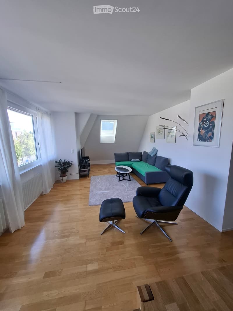 Modern living room with wooden flooring, large windows, a green sofa, a black leather chair, a coffee table, a small plant, and artwork on the walls.