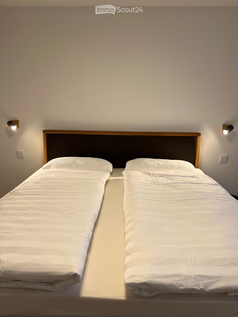 A bedroom with two single beds, each with white bedding and a dark headboard, placed side by side against a plain wall.
