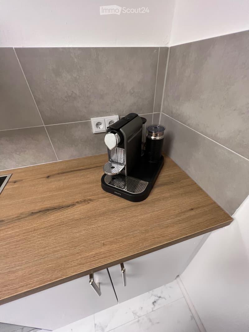 A modern kitchen corner with a wooden countertop and a black coffee machine placed on it. The walls are tiled in a grey pattern, and the floor is marble.