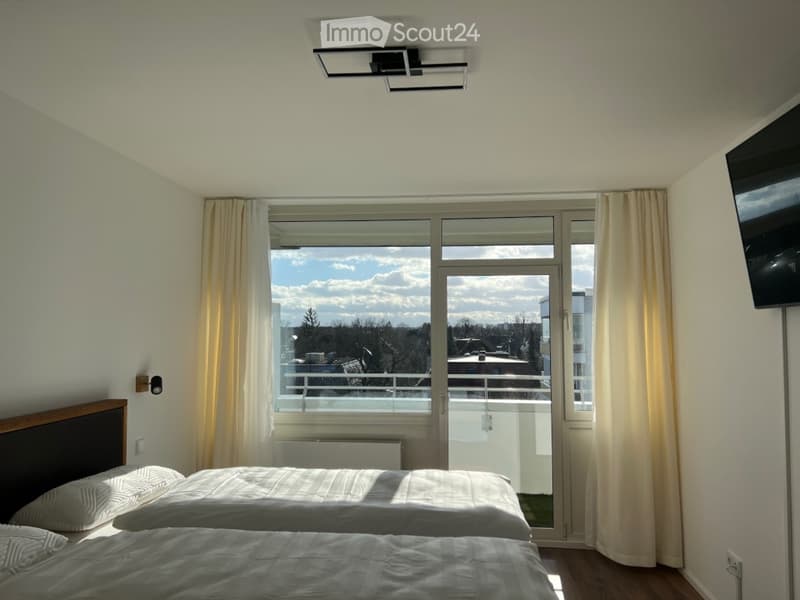 Bright bedroom with two single beds, large window with balcony access, and a wall-mounted TV.