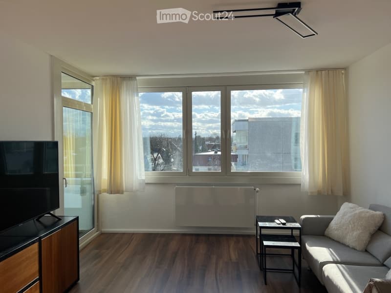 Modern living room with large windows offering a view, a grey sofa with a white throw pillow, a coffee table, a television on a wooden stand, and a radiator below the windows.