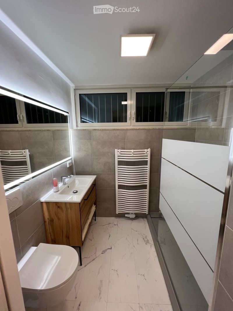 Modern bathroom with marble flooring, a wooden vanity with a sink, a toilet, a towel rack, and a large mirror with built-in lighting.