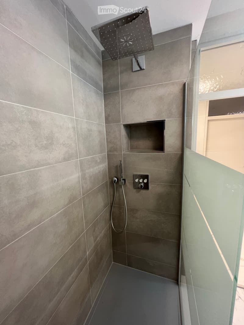 Modern shower area with grey tiles, a large overhead showerhead, a control panel, and a built-in shelf.