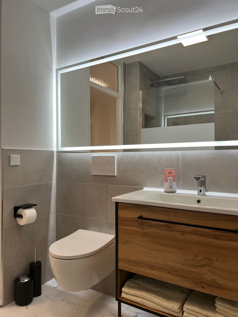 Modern bathroom with a sleek sink, wooden cabinet, large mirror with built-in lighting, toilet, and neatly stacked towels.